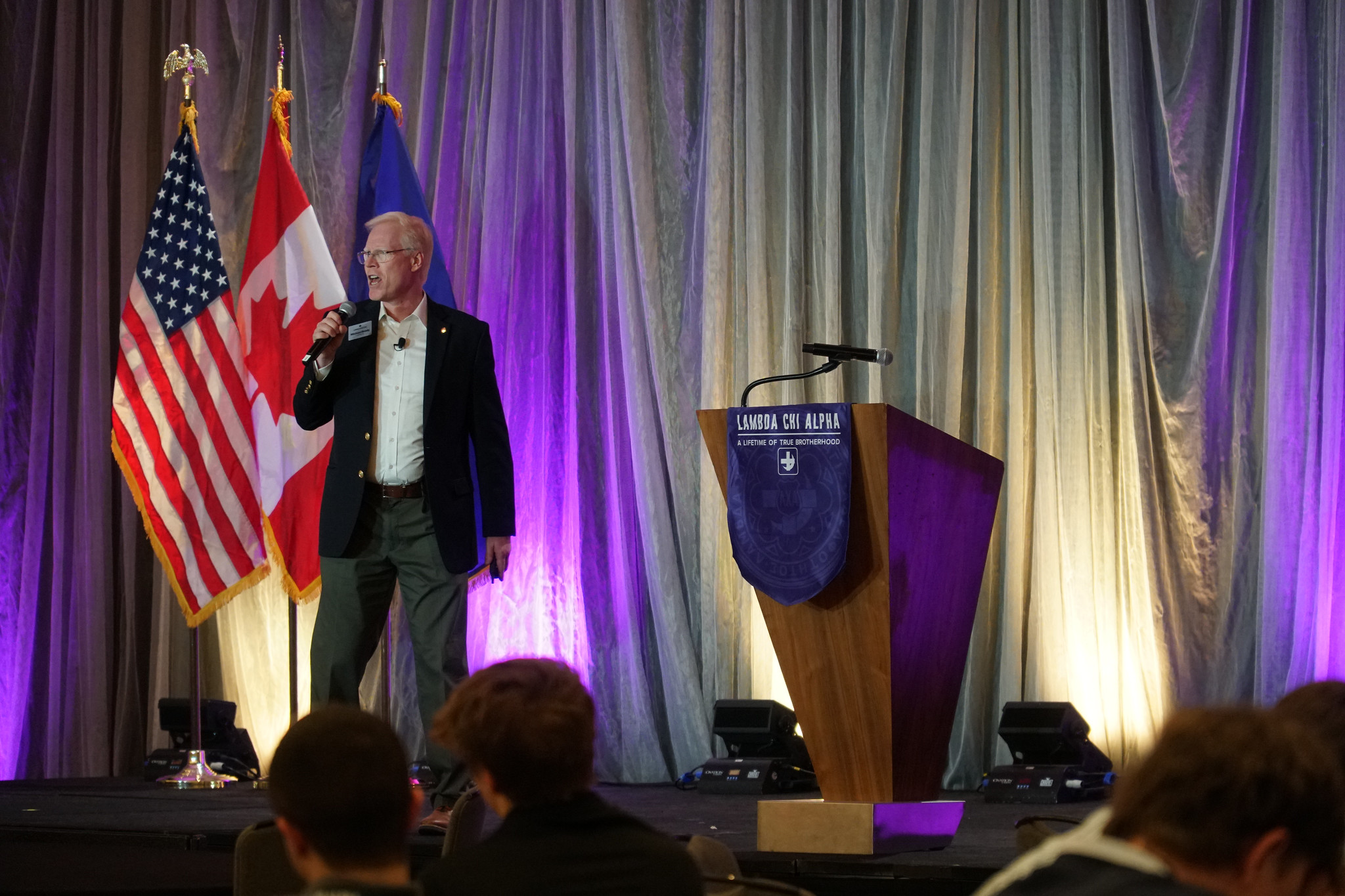 Michael Brady speaking on stage at a Lambda Chi Alpha leadership event, standing beside a podium with fraternity branding and U.S. and Canadian flags in the background.